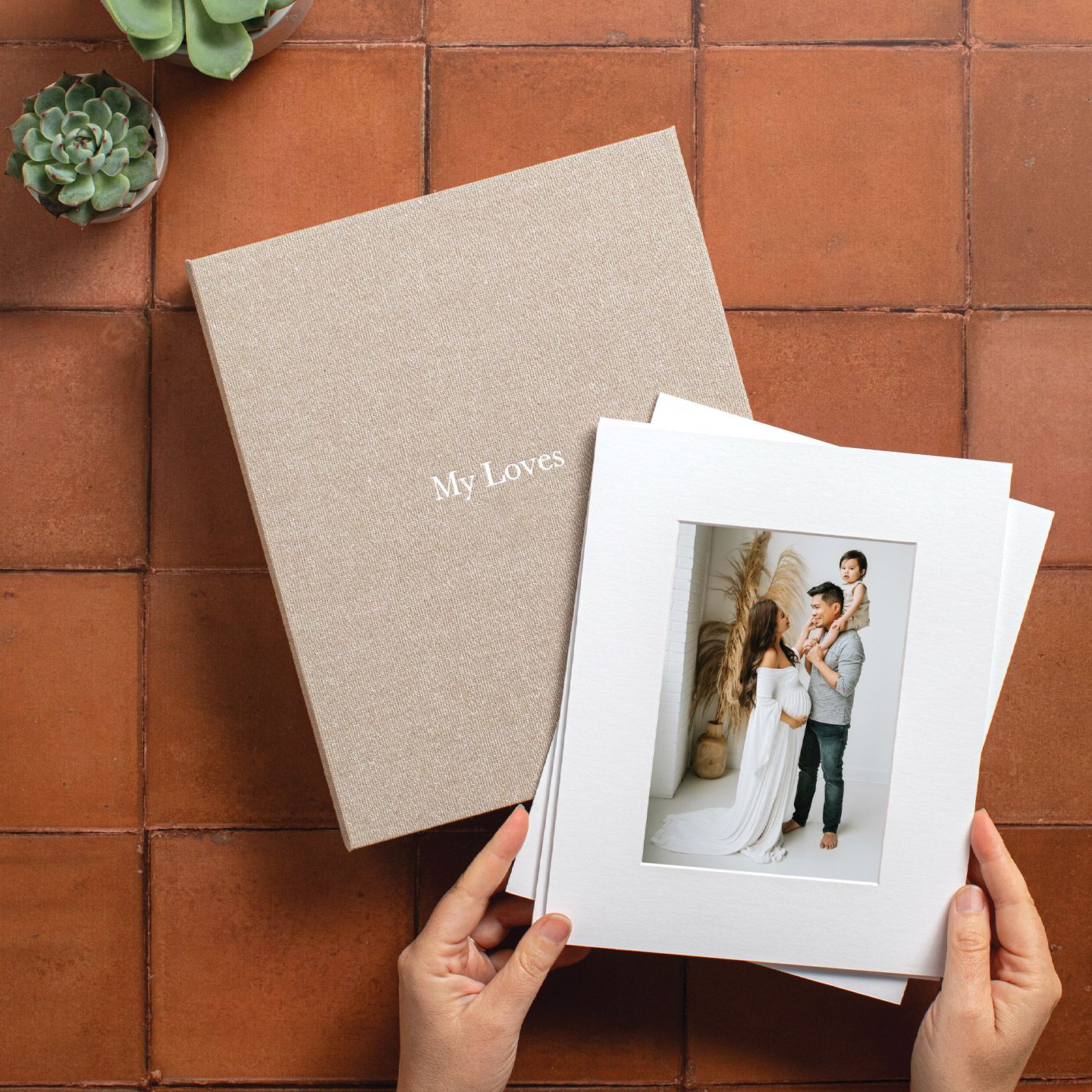 Hands holding a white matted print of a maternity portrait, beside a linen album cover embossed “My Loves” on terracotta tile