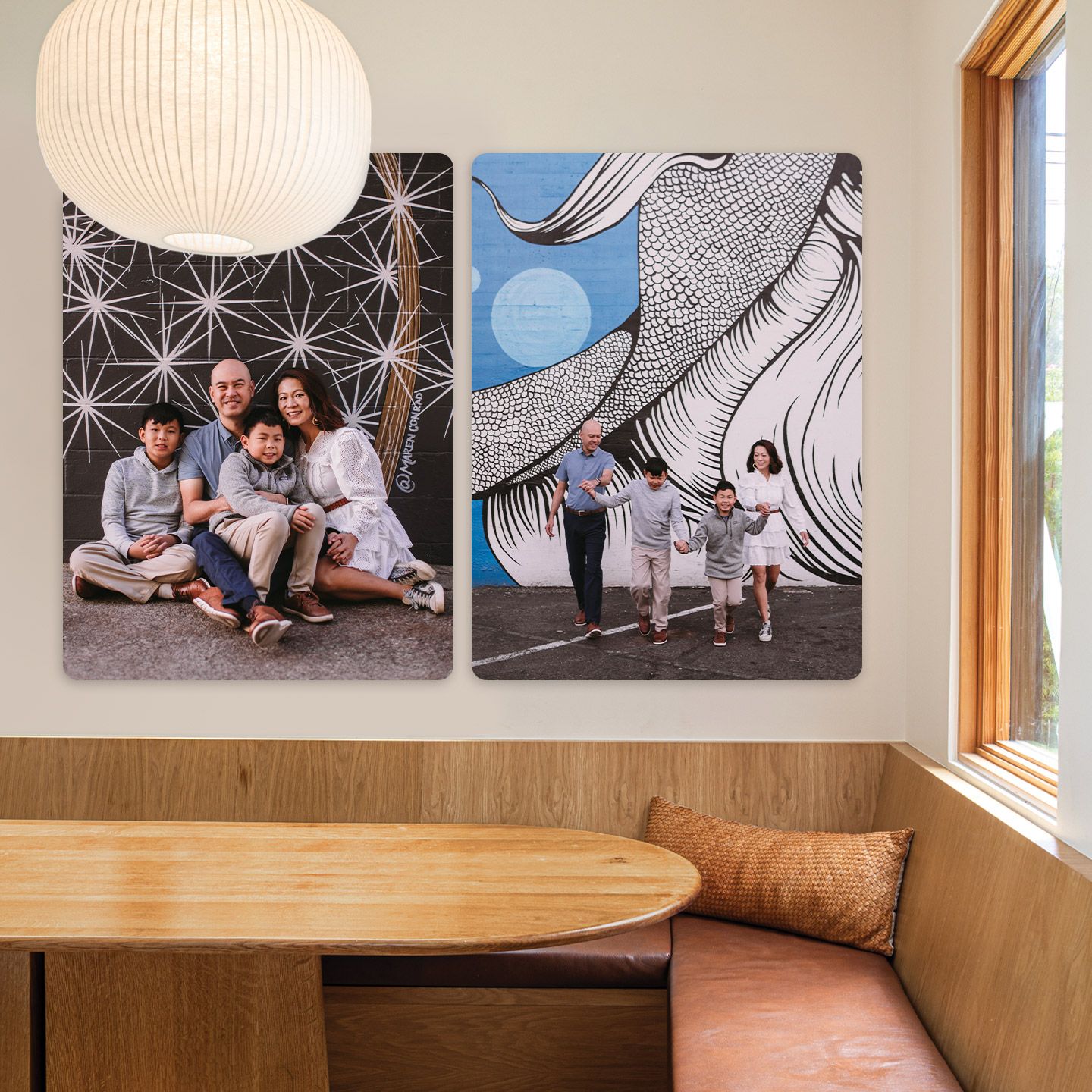 Two metal prints of a family photographed against painted murals, hanging above a wooden banquette in a sunlit dining nook