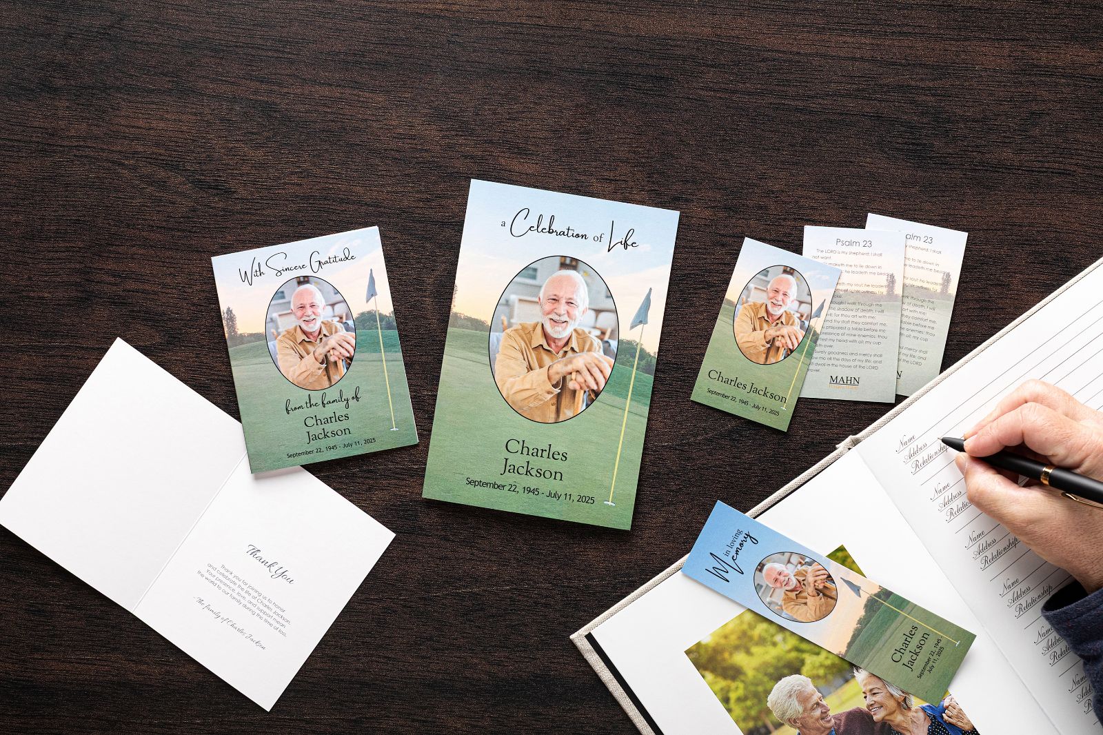 A matching set of memorial products laid out on a dark wood table — service programs, prayer cards, thank you cards, and bookmarks — while a guest signs the registration book