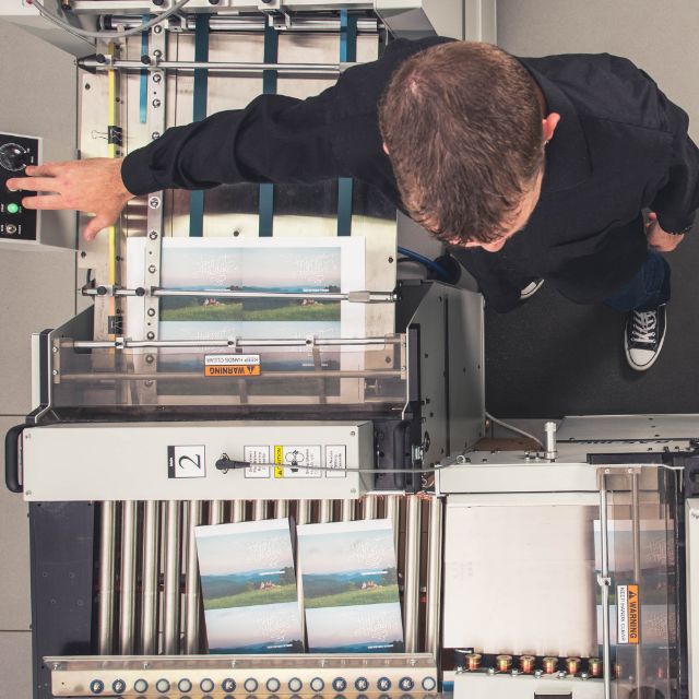 Overhead view of a Miller's press operator running a printing press, freshly printed cards stacked at the delivery end